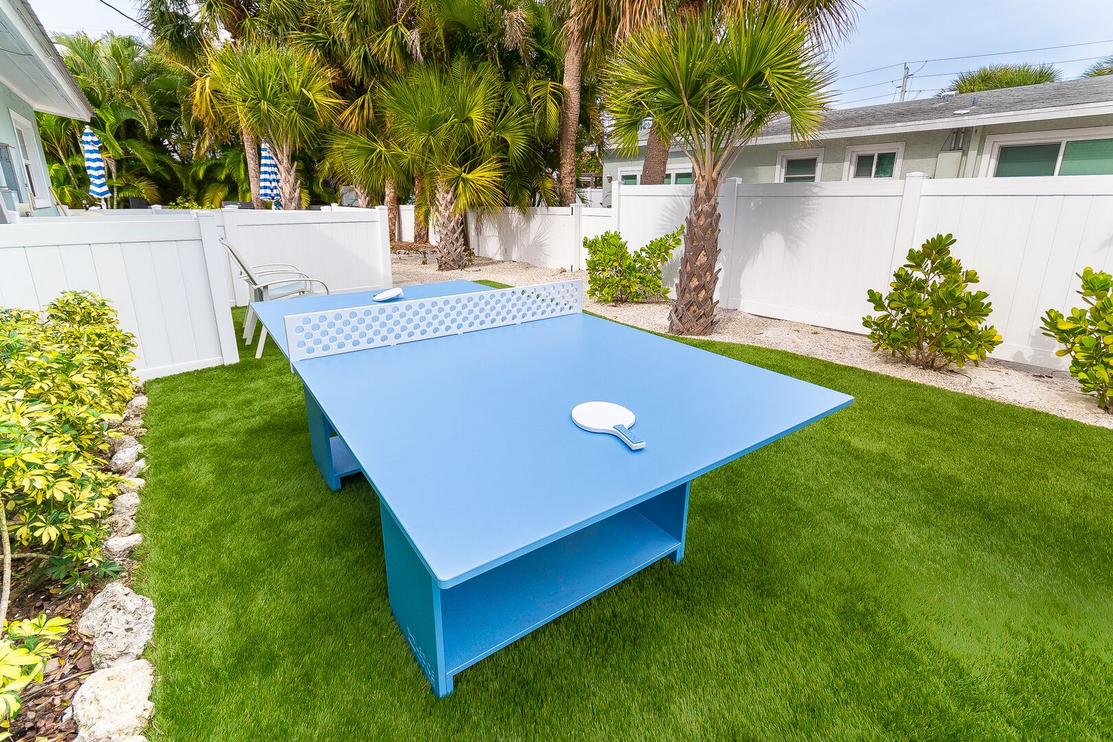 Une table de ping-pong bleue avec une raquette au centre, entourée d’herbe verte et de palmiers.