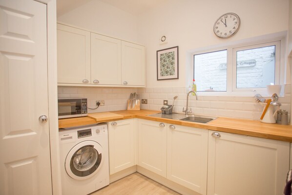 A compact kitchen with white cabinets, a washing machine, a microwave, and a sink. - Foresters Cottage (Worthing)