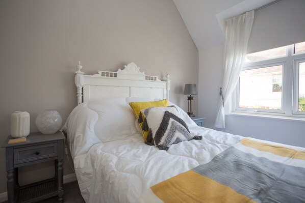 A bedroom with a white bed, a nightstand, a lamp, and a window with curtains. - Foresters Cottage (Worthing)
