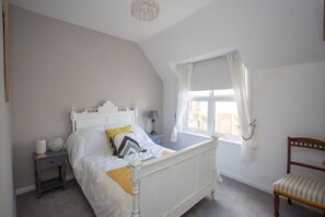 Une chambre à coucher avec un lit blanc, une table de chevet, une lampe, une chaise et une fenêtre avec des rideaux.