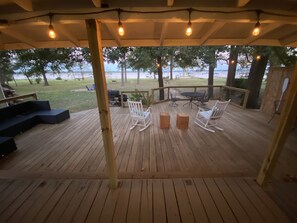 Una terraza de madera con mobiliario exterior, como sillas y una mesa, con vistas a una zona verde con árboles y un cuerpo de agua.