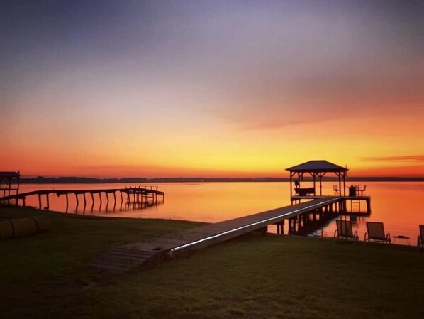 Vista de un atardecer con un muelle que se adentra en agua tranquila, un pabellón y una zona ajardinada con sillas.