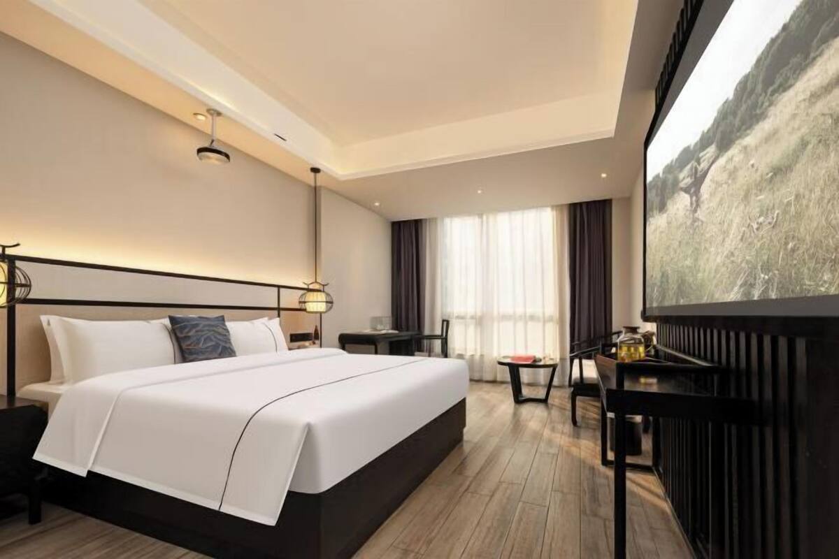 A hotel room with a large bed, a desk, a chair, a small table, and a large window with a view.