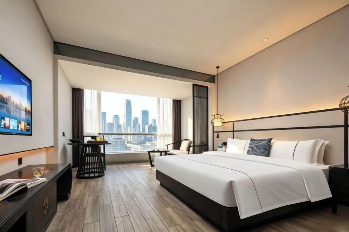 A modern hotel room with a large bed, a desk, a TV, and a city view.