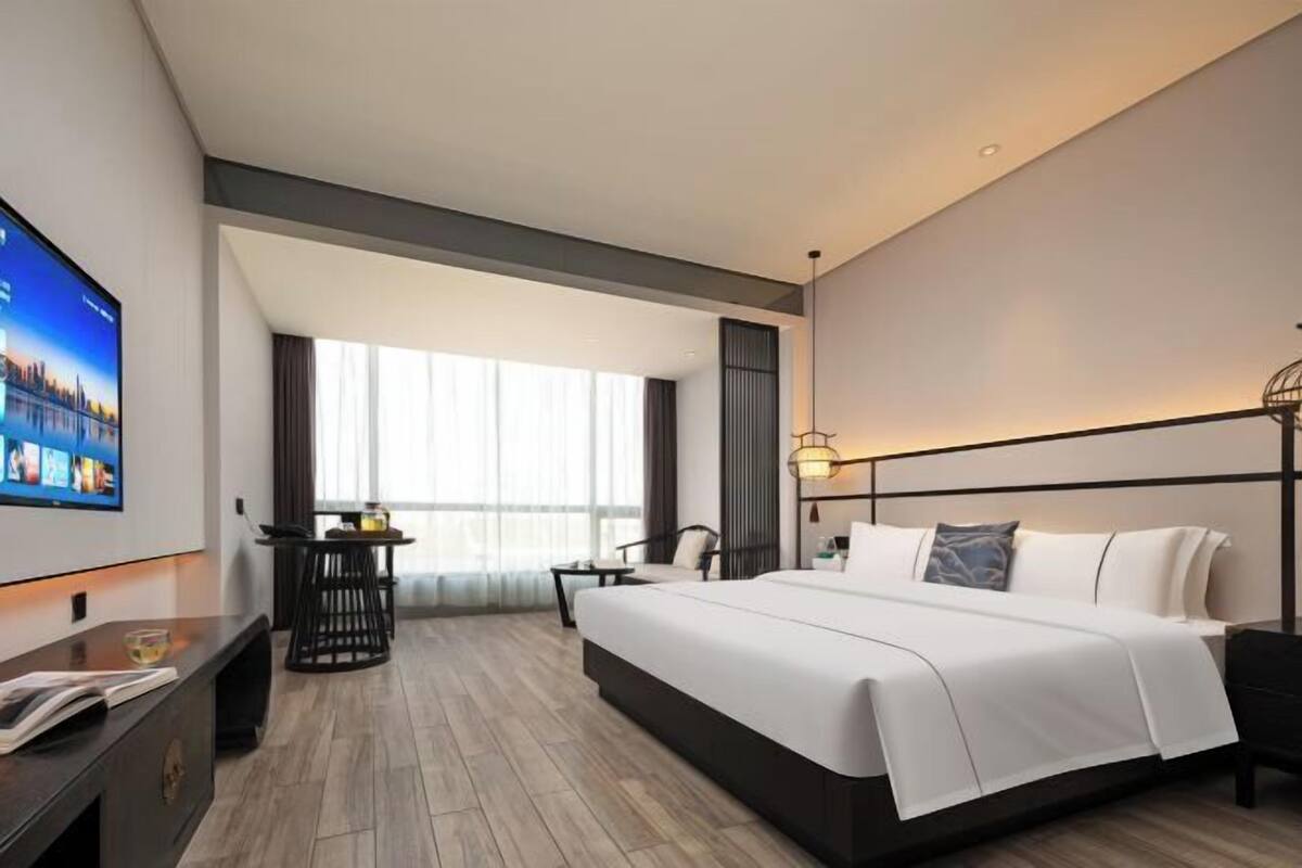 A modern hotel room with a large bed, a flat-screen TV, a desk, and a window with curtains.