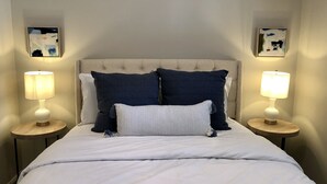 A neatly made bed with a white comforter and two pillows, flanked by two bedside tables with lamps, in a room with a tufted headboard and framed artwork on the walls.