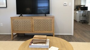 A living room with a flat-screen TV on a wooden cabinet, a round coffee table with books, and a kitchen in the background.