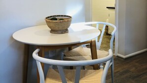 A dining area with a round table, two white chairs with woven seats, a framed picture on the wall, and a basket on the table.
