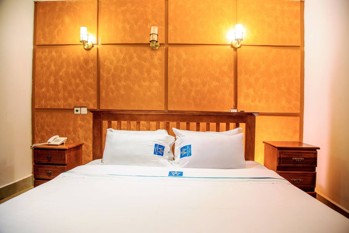 Ein Hotelzimmer mit einem hölzernen Kopfteil, zwei Nachttischen, einem weißen Bett mit blauer Beschriftung und Wandlampen.