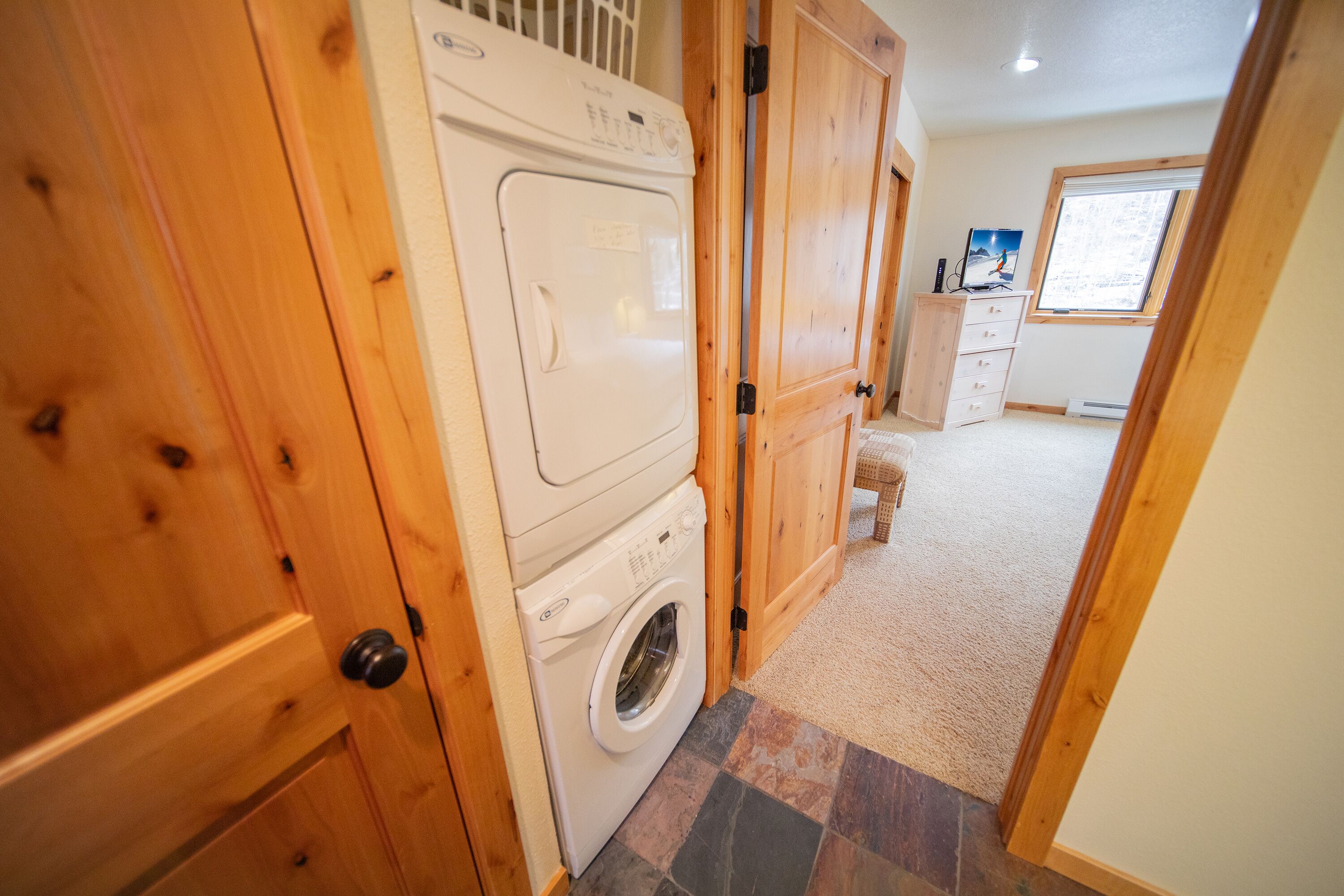 A laundry area with a front-loading washer and dryer, wooden doors, and a view of a bedroom with a window.