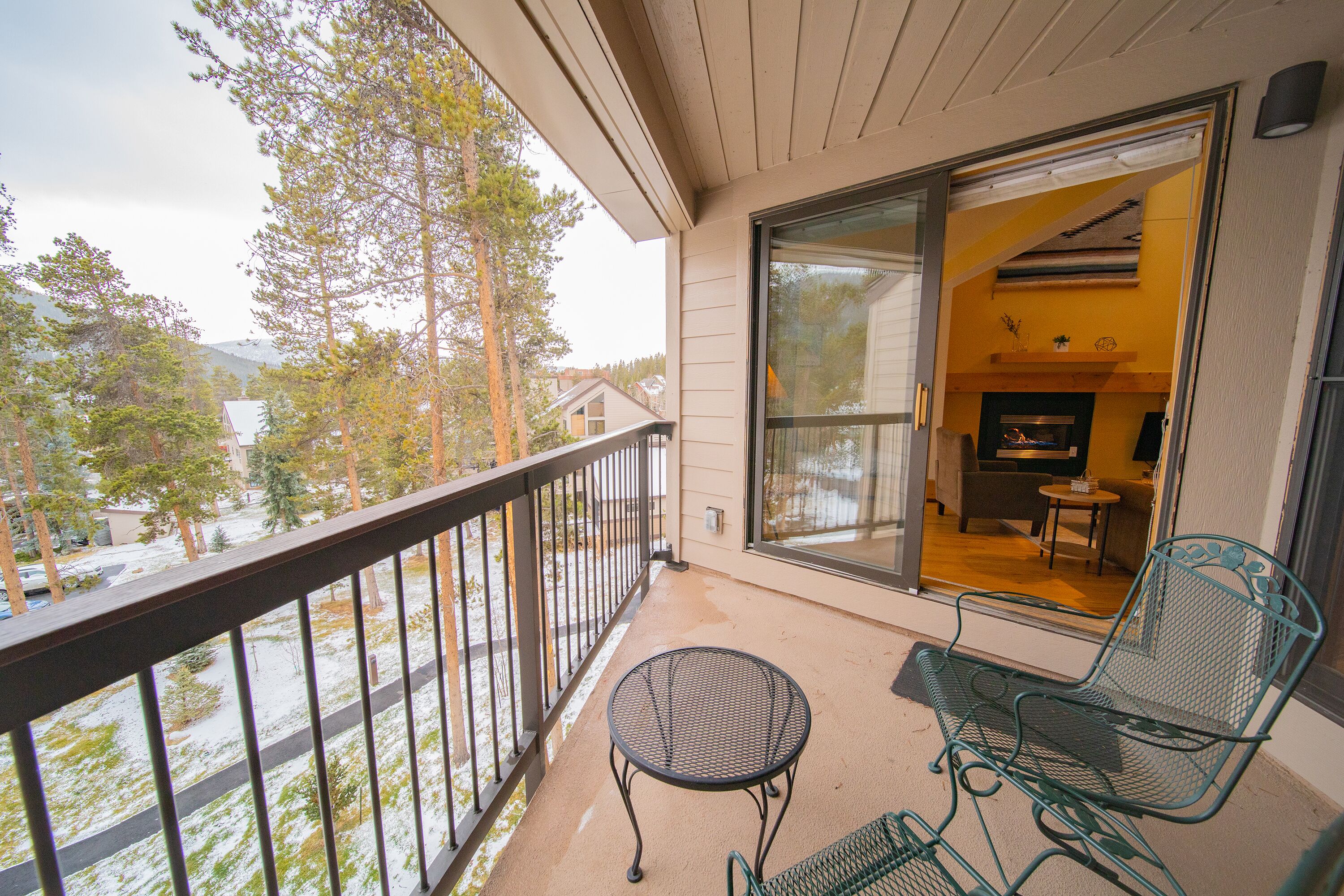 A balcony with a metal table and chairs, a glass door leading inside, and a view of trees and a road.