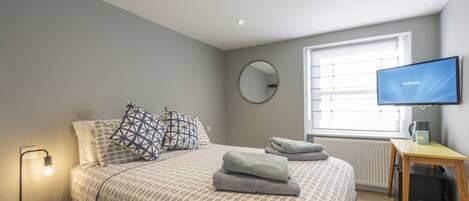 A modern bedroom with a bed, a bedside lamp, a television, a mirror, and a window with blinds.