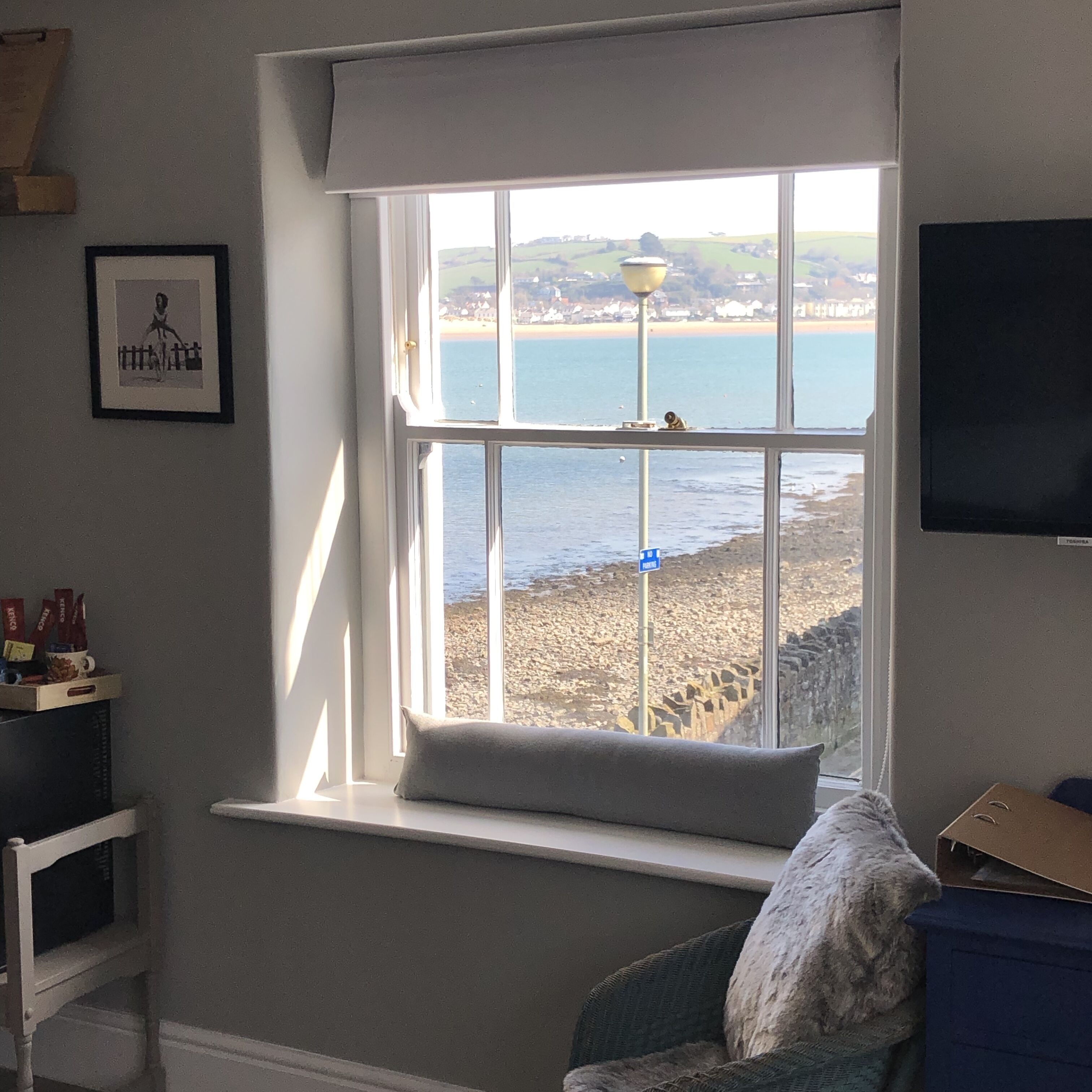 A room with a large window overlooking a beach, a framed picture on the wall, and a television on the right.