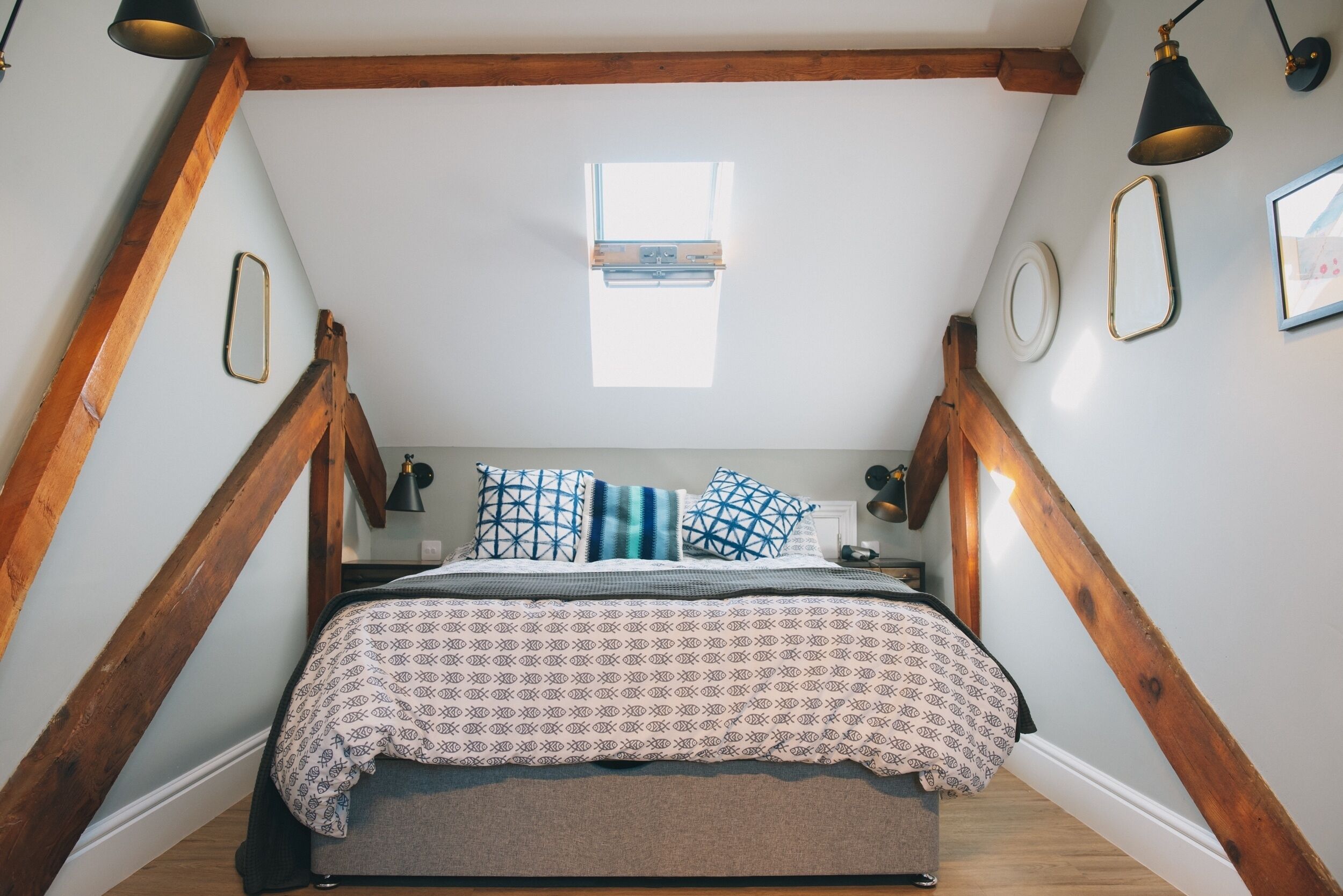 Een slaapkamer met een schuine plafond, houten balken, een bed met beddengoed met een patroon en een klein raam.