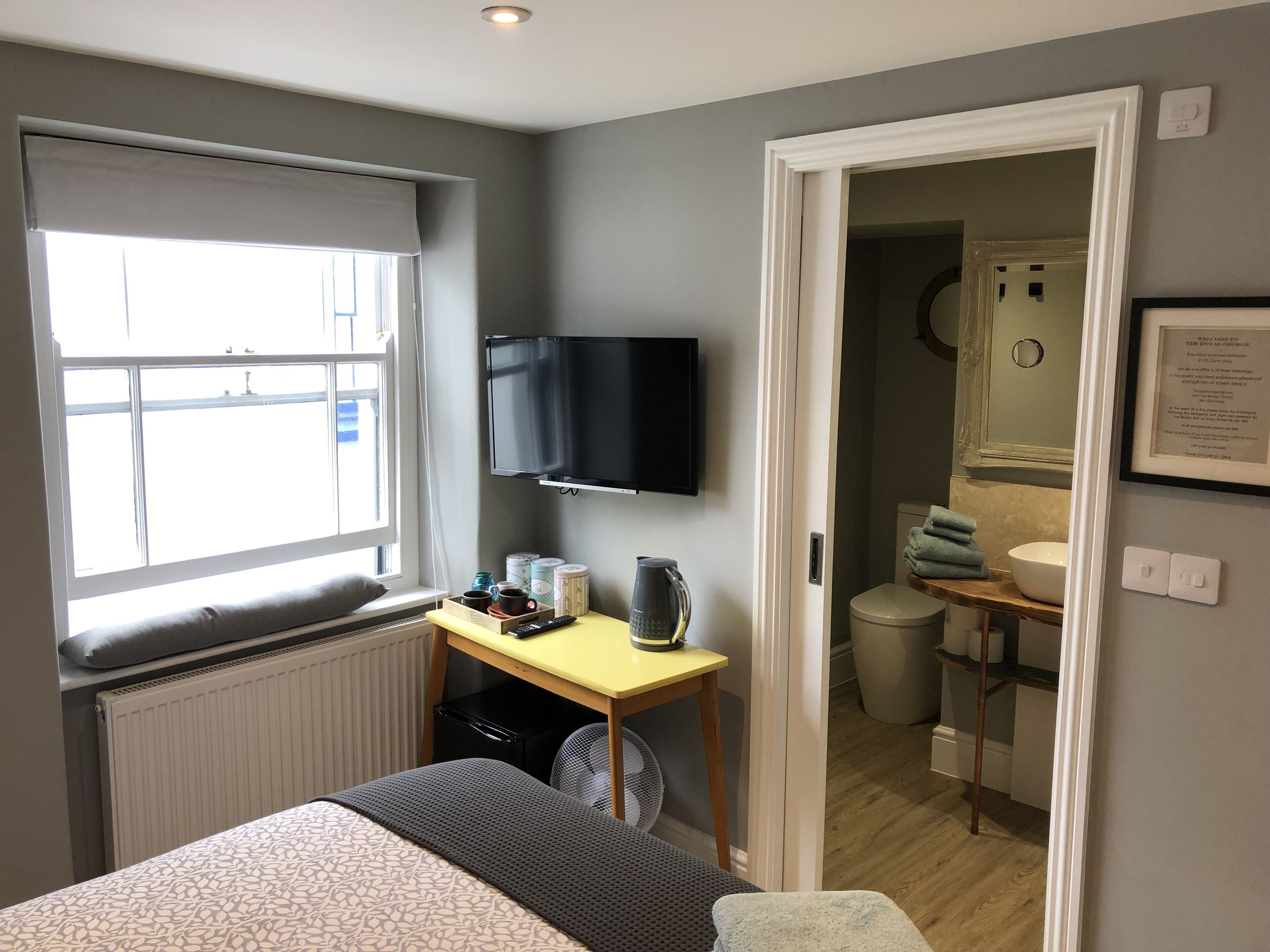Ein Hotelzimmer mit Bett, Schreibtisch mit Fernseher, Fenster und einem durchgegangene Tür erkennbaren Badezimmer.