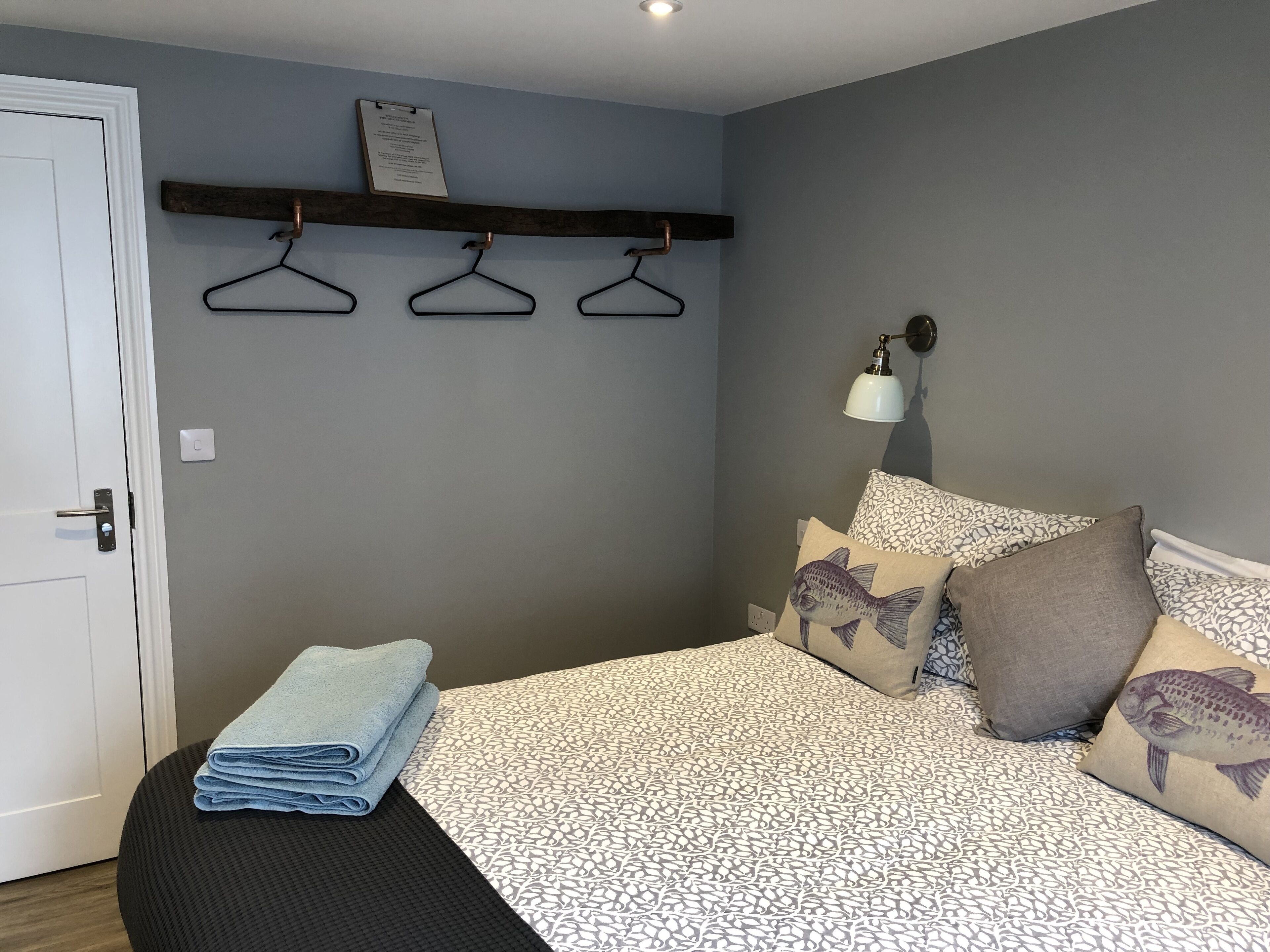 A bedroom with a bed, a bedside table, a wall-mounted shelf with hangers, a wall lamp, and a door.