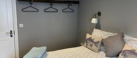 A bedroom with a bed, a bedside table, a wall-mounted shelf with hangers, a wall lamp, and a door.