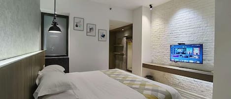 A hotel room with a bed, a TV mounted on a brick wall, a pendant light, and a door leading to a bathroom.