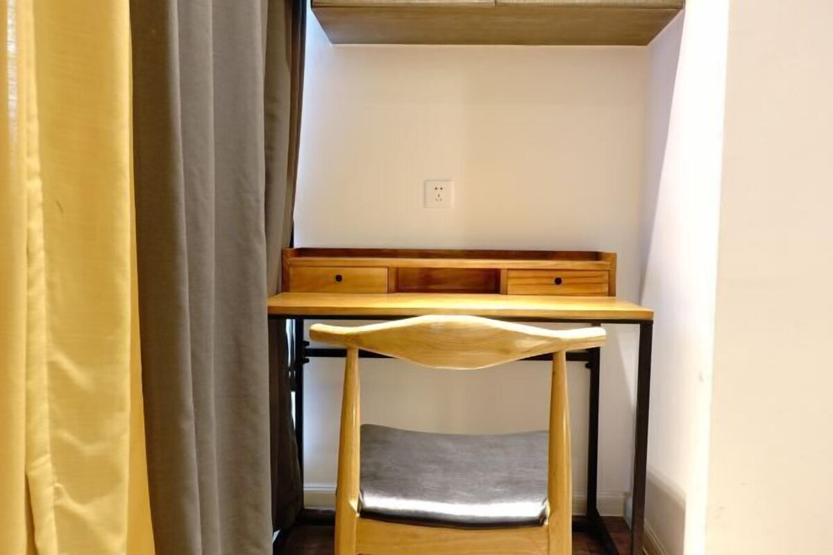A small, narrow room with a wooden desk and chair, a white wall, and a power outlet.