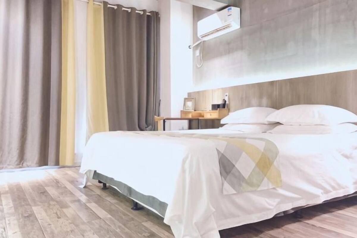 A hotel room with a bed, curtains, an air conditioning unit, and a wooden floor.