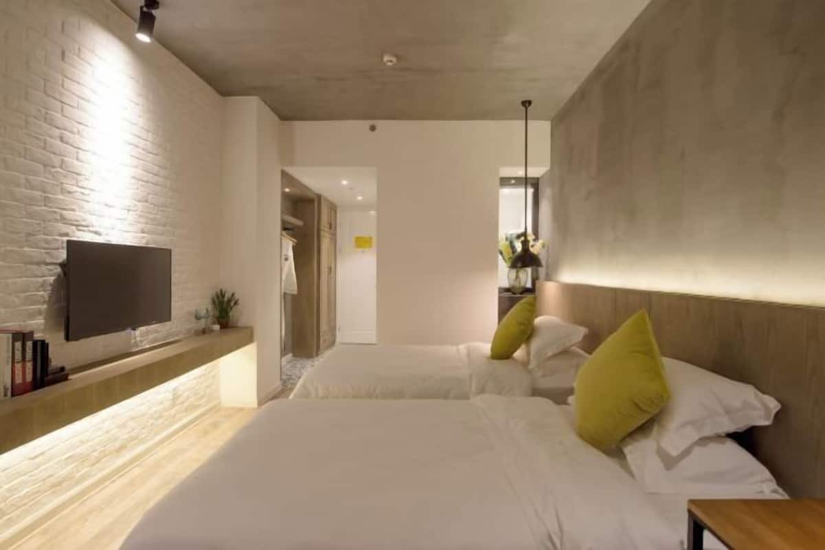 A modern hotel room with a brick accent wall, a flat-screen TV, two beds with white bedding and green pillows, a wooden bedside table, and a small plant.