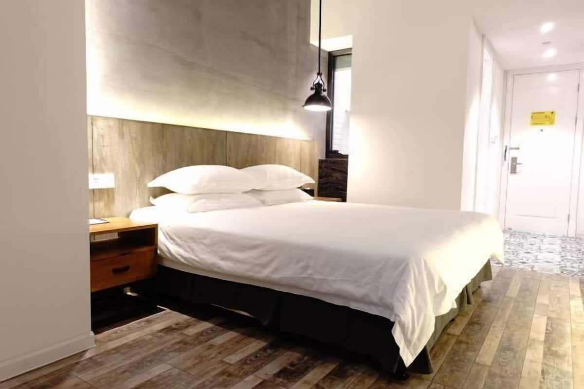 A modern hotel room with a wooden headboard, a single bed with white bedding, a nightstand, a lamp, and a wooden floor.