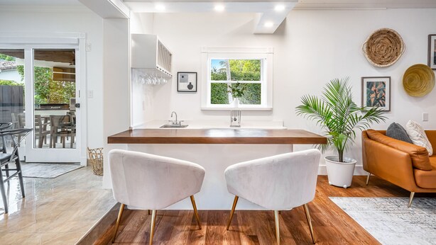 A modern kitchen with a central island, white cabinetry, and a large window. There are two white chairs in front of the island, a brown leather sofa, and a potted plant. - 5-3 1/2 Aventura-Heated Pool-Gas Grill-Coverd Patio (North Miami Beach)