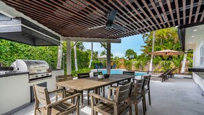 An outdoor patio with a wooden table and chairs, a grill, and a pool in the background. - 5-3 1/2 Aventura-Heated Pool-Gas Grill-Coverd Patio (North Miami Beach)