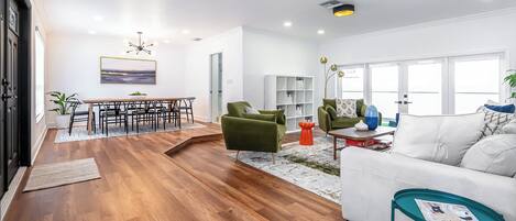 A spacious living room with a wooden floor, a white sofa, green armchairs, and a dining area with a wooden table and chairs.
