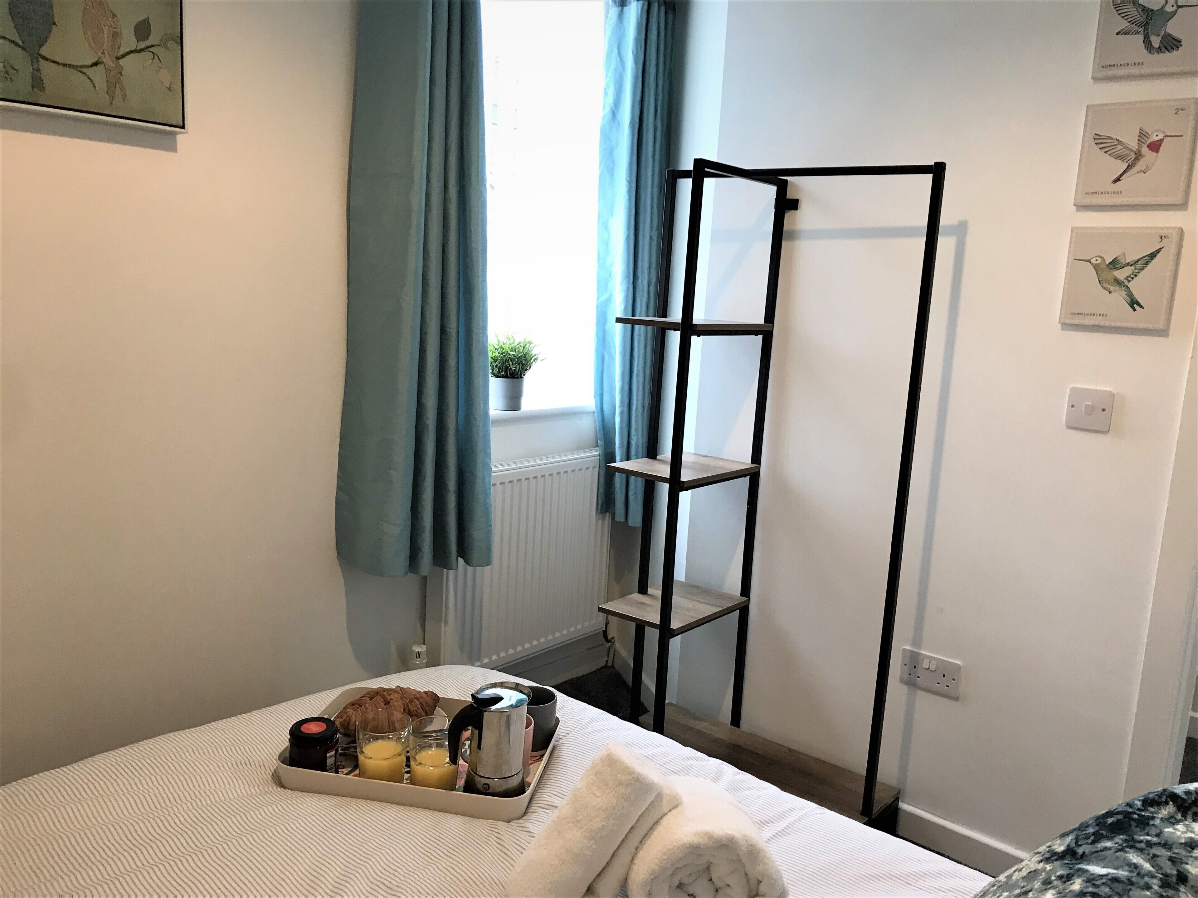 A neatly made bed with a tray of breakfast items, a black metal shelf unit, and a window with blue curtains.