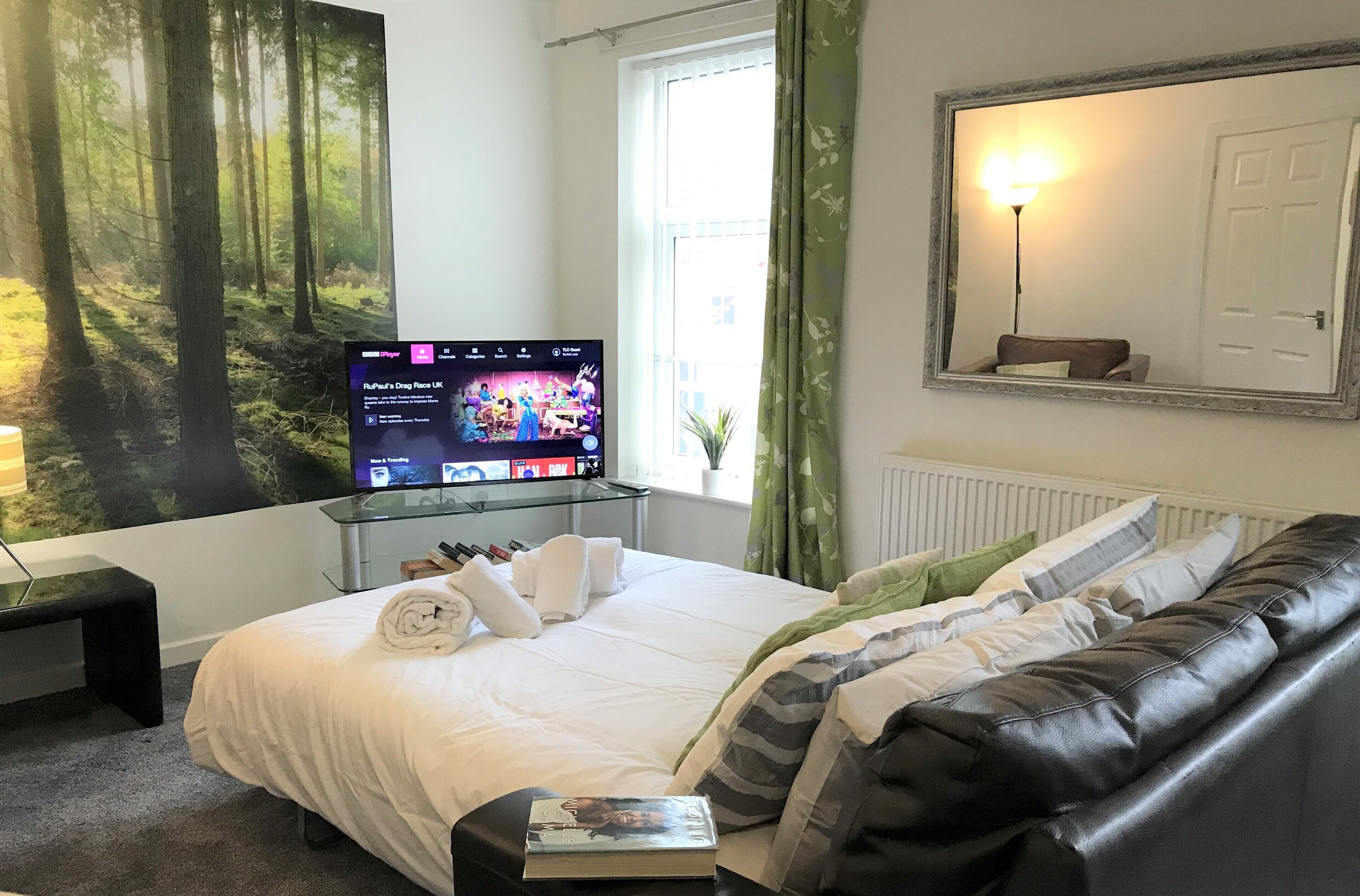 A bedroom with a bed, a TV on a glass table, a mirror, and a window with curtains.