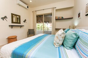 Un dormitorio con una cama, almohadas, una silla, una estantería con libros, un espejo y una ventana con cortinas.