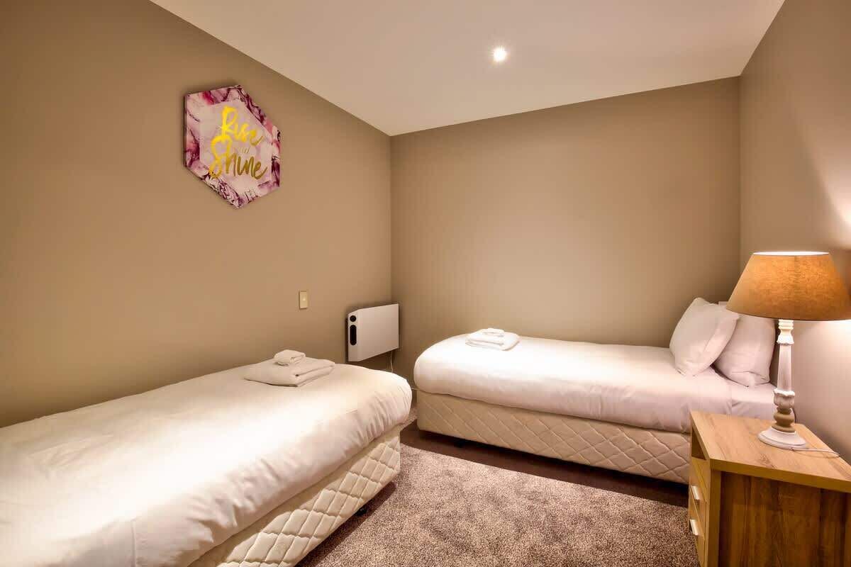 Een kamer met twee eenpersoonsbedden, een houten nachtkastje met een lamp, een wandradiator en een decoratief bord.