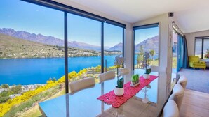 A dining area with a table set for four, large windows offering a view of a lake and mountains, and a bright yellow chair in the background. - Ultimate Heights Queenstown Jaw dropping views (Queenstown)