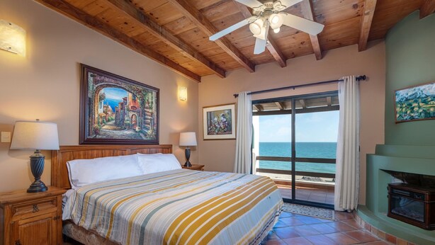 A bedroom with a large bed, wooden headboard, a painting of a coastal scene, a fireplace, and a view of the sea through a sliding door. - Paradise Villa 20 by Kivoya (Puerto Peñasco)