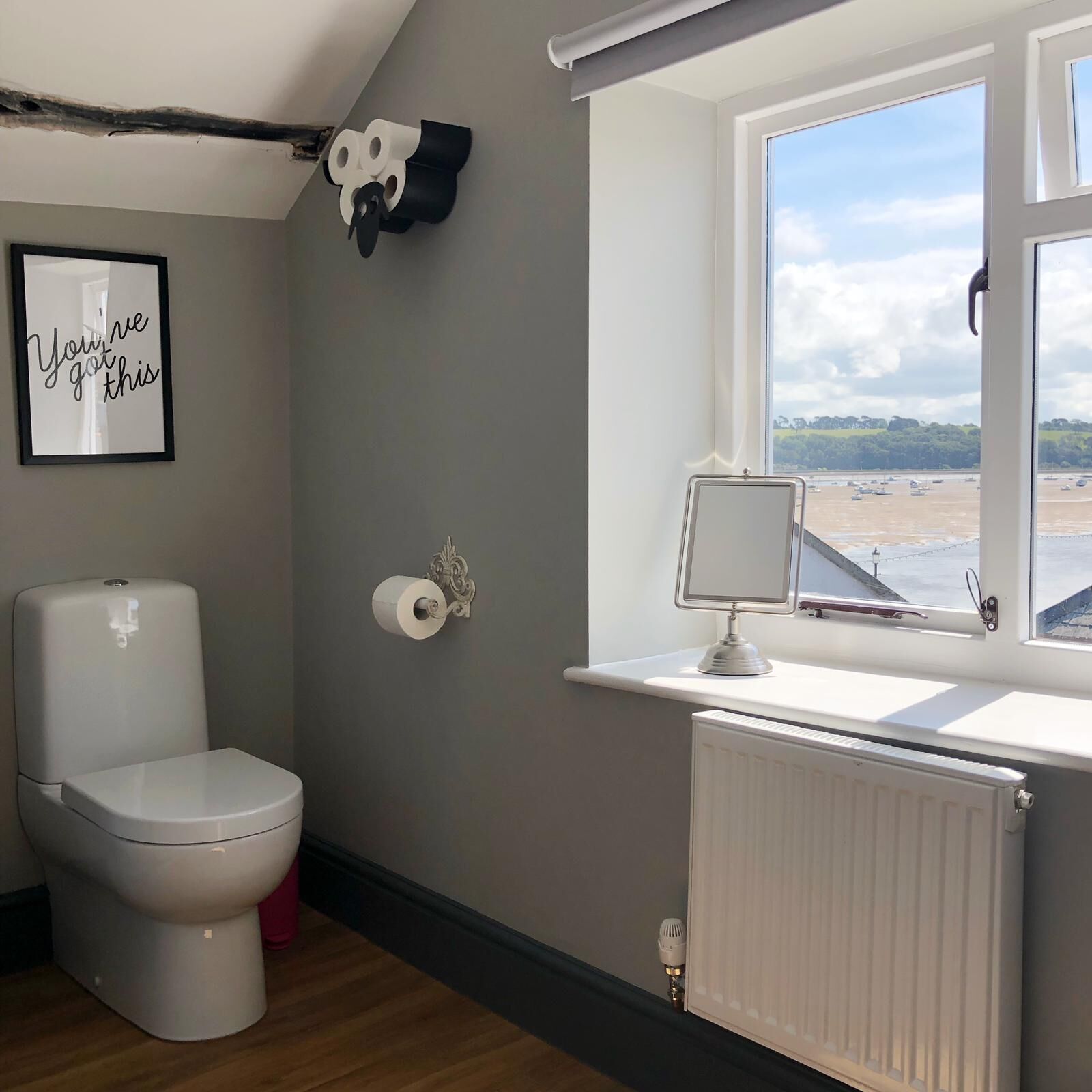 A bathroom with a toilet, a toilet paper holder, a radiator, and a window with a view of a beach and boats.