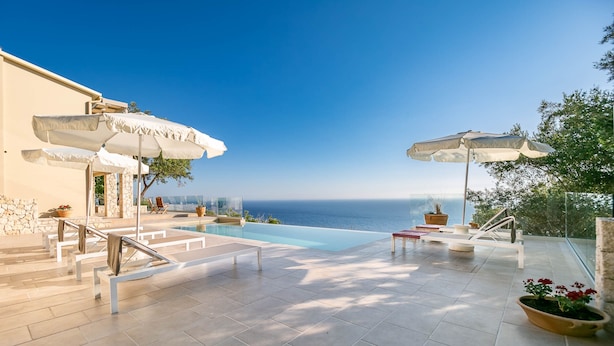 A pool area with a clear view of the sea, outdoor seating, and umbrellas. - Ocean View - Luxury Villa Ethra (Corfu)