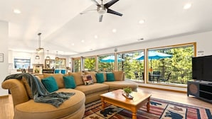 A modern living room with a sectional sofa, a coffee table, a ceiling fan, and large windows with outdoor views. - Serene Tahoe Escape | HotTub | Arcade | EV Charger (South Lake Tahoe)