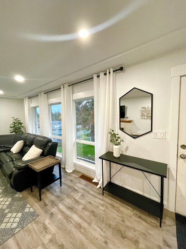 A modern living room with a black leather sofa, a wooden side table, a mirror, and a window with white curtains.