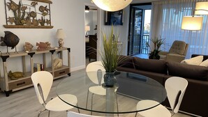 A modern living room with a glass dining table surrounded by white chairs, a brown sofa, and a large window with curtains. - Vanderbilt Beach area with Bay views from Lanai. (Naples)