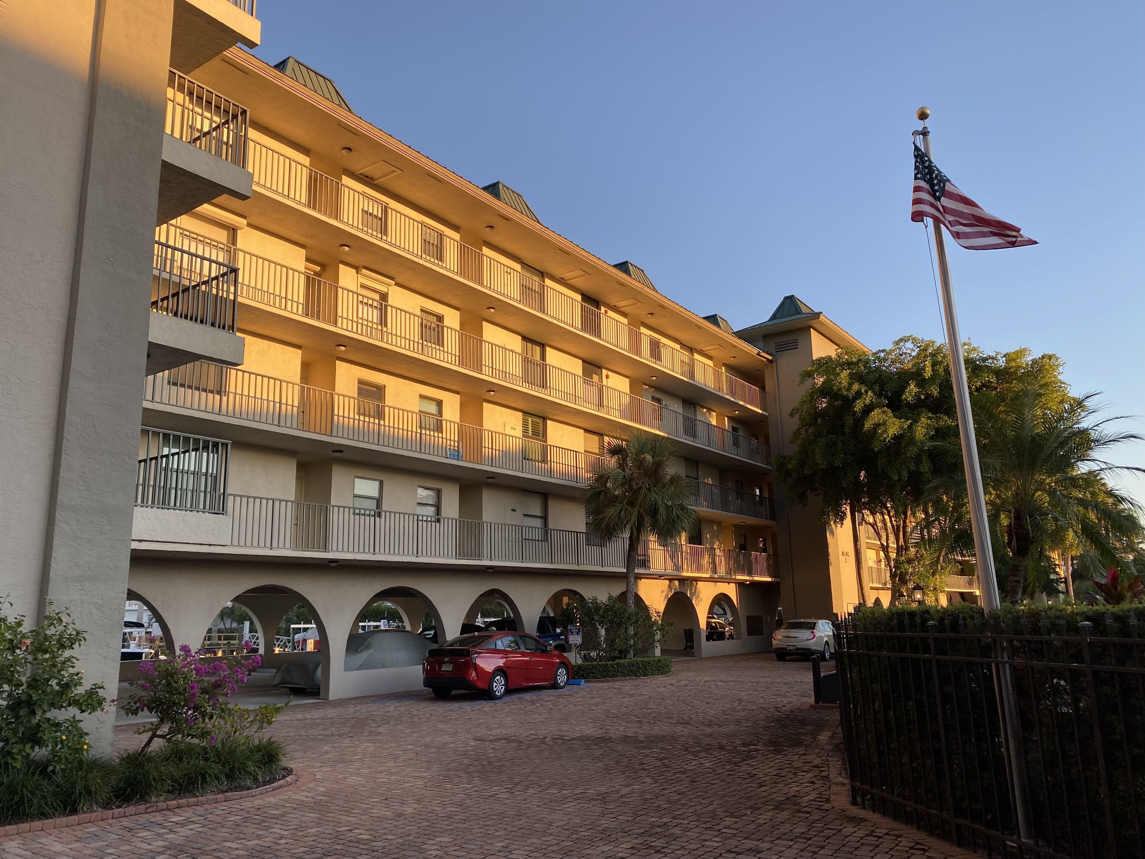 A multi-story hotel with balconies, a flagpole with an American flag, and a paved driveway with cars.