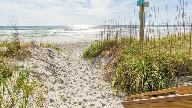 Un sentier de sable qui longe la plage et mène à l’océan, avec une promenade en bois et un panneau de signalisation.