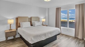 A bedroom with a large bed, two bedside tables with lamps, a window with curtains, and a view of the beach. - Beach Front Condo at the Avalon Club (Clearwater)