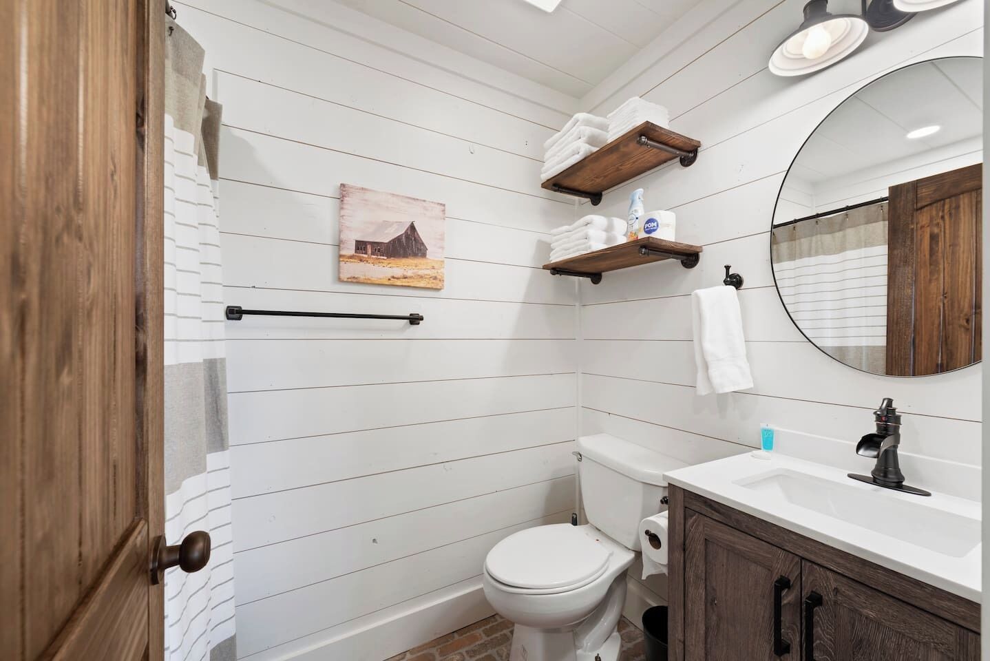 Un baño con pared de azulejos blancos, puerta de madera, inodoro, lavabo con espejo y un perchero para toallas.