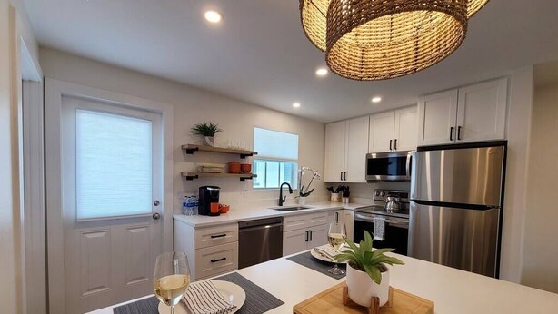 Una cocina moderna con armarios blancos, electrodomésticos de acero inoxidable, una isla central con una copa de vino blanco y una lámpara colgante de fibras naturales.