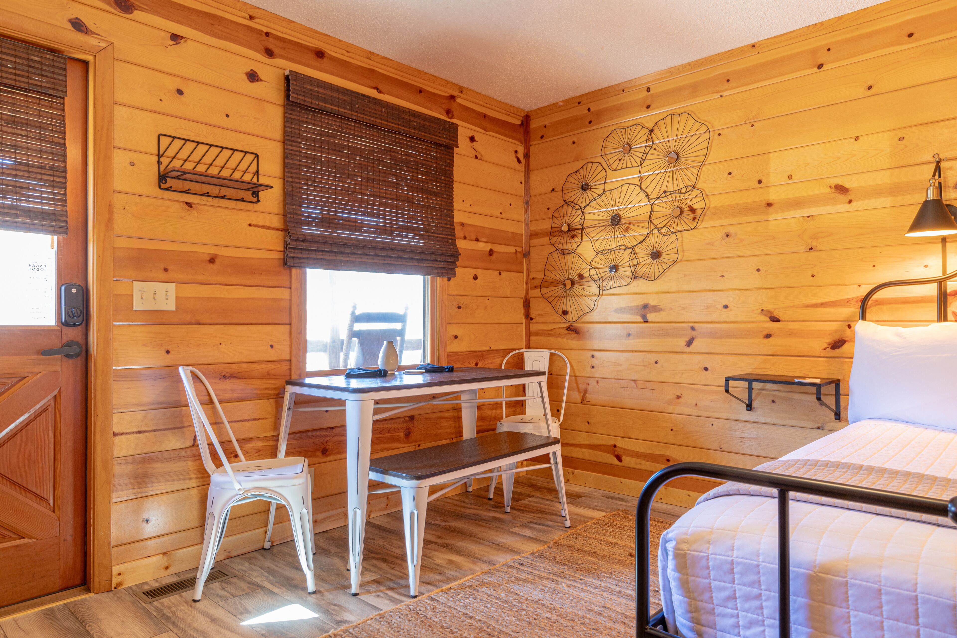 A wooden cabin room with a bed, a desk with a chair, a small table, and a wall-mounted shelf.