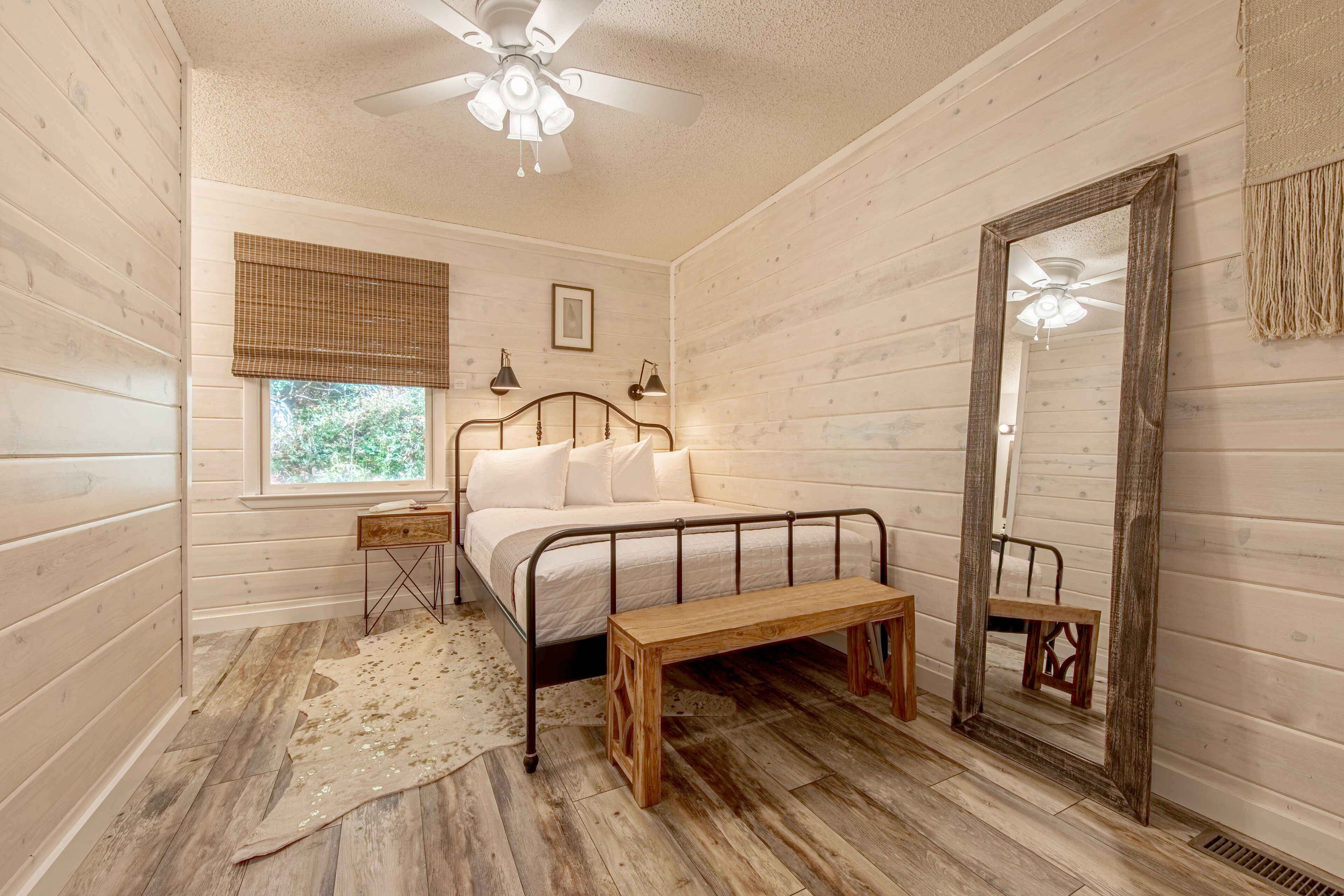 A bedroom with a wooden floor, a bed with a metal frame, a wooden bench, a ceiling fan, a window with blinds, and a mirror.
