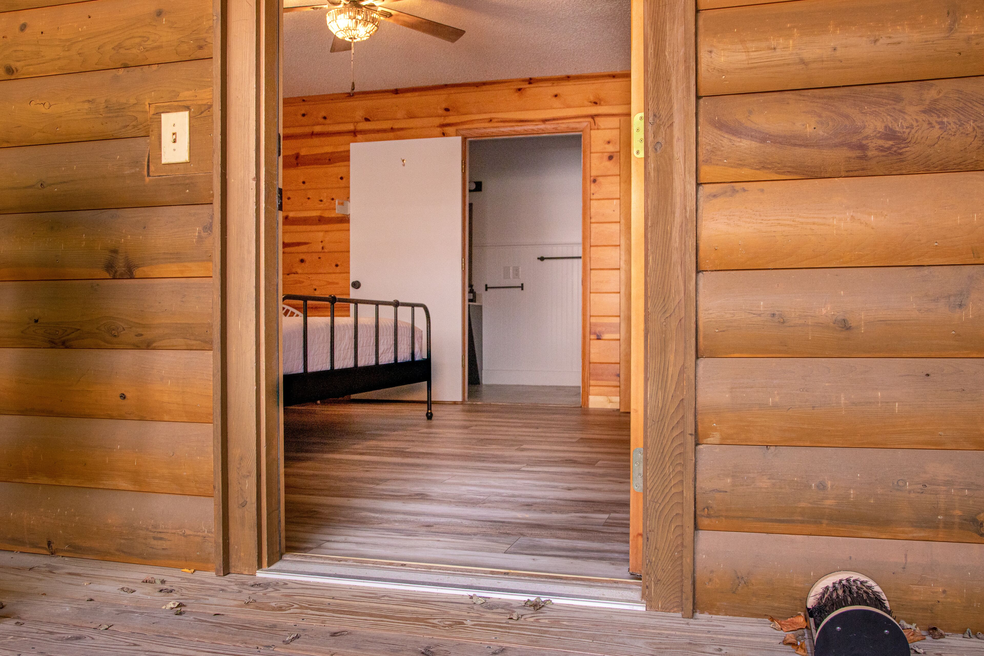 L’intérieur d’un chalet en bois, avec un lit au premier plan, une porte donnant sur une autre pièce et un ventilateur de plafond.