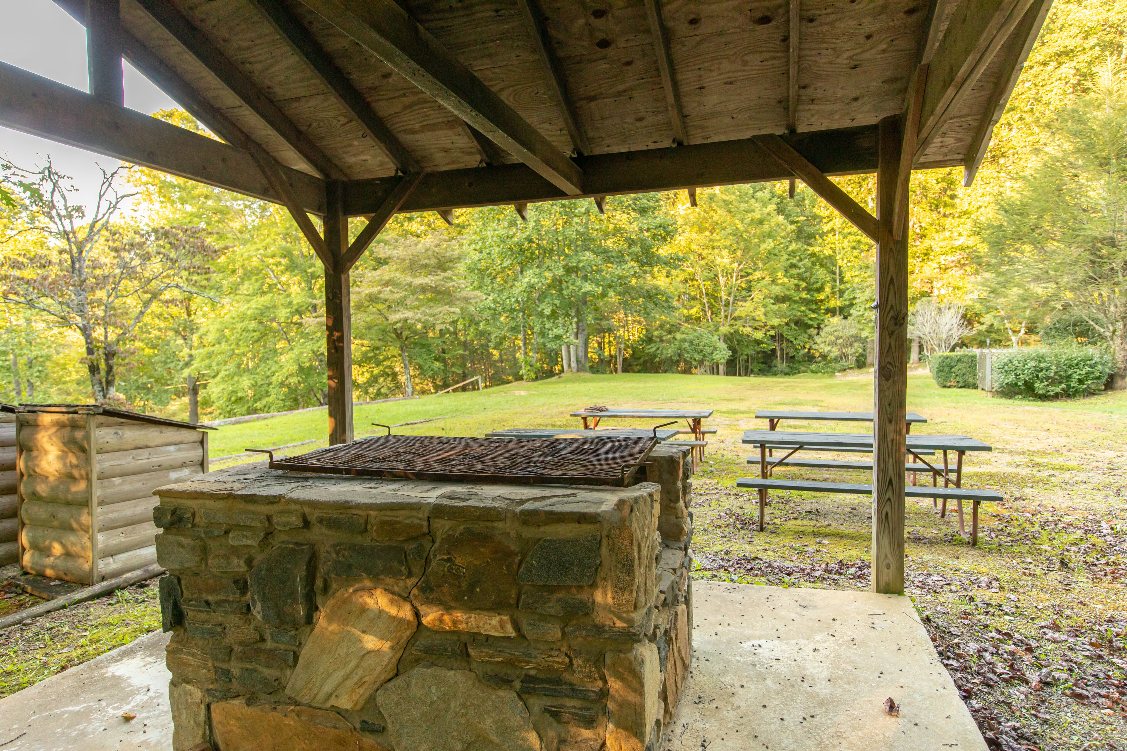 A covered outdoor area with a stone barbecue and picnic tables, surrounded by a park with trees and a grassy lawn.