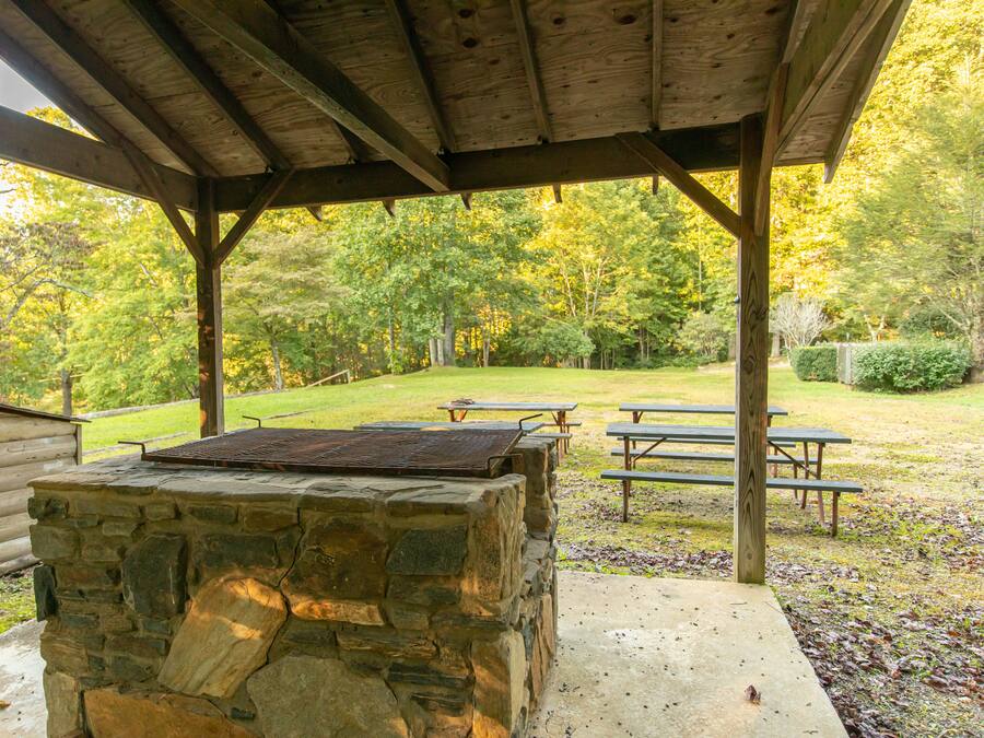 A covered outdoor area with a stone barbecue and picnic tables, surrounded by a park with trees and a grassy lawn.